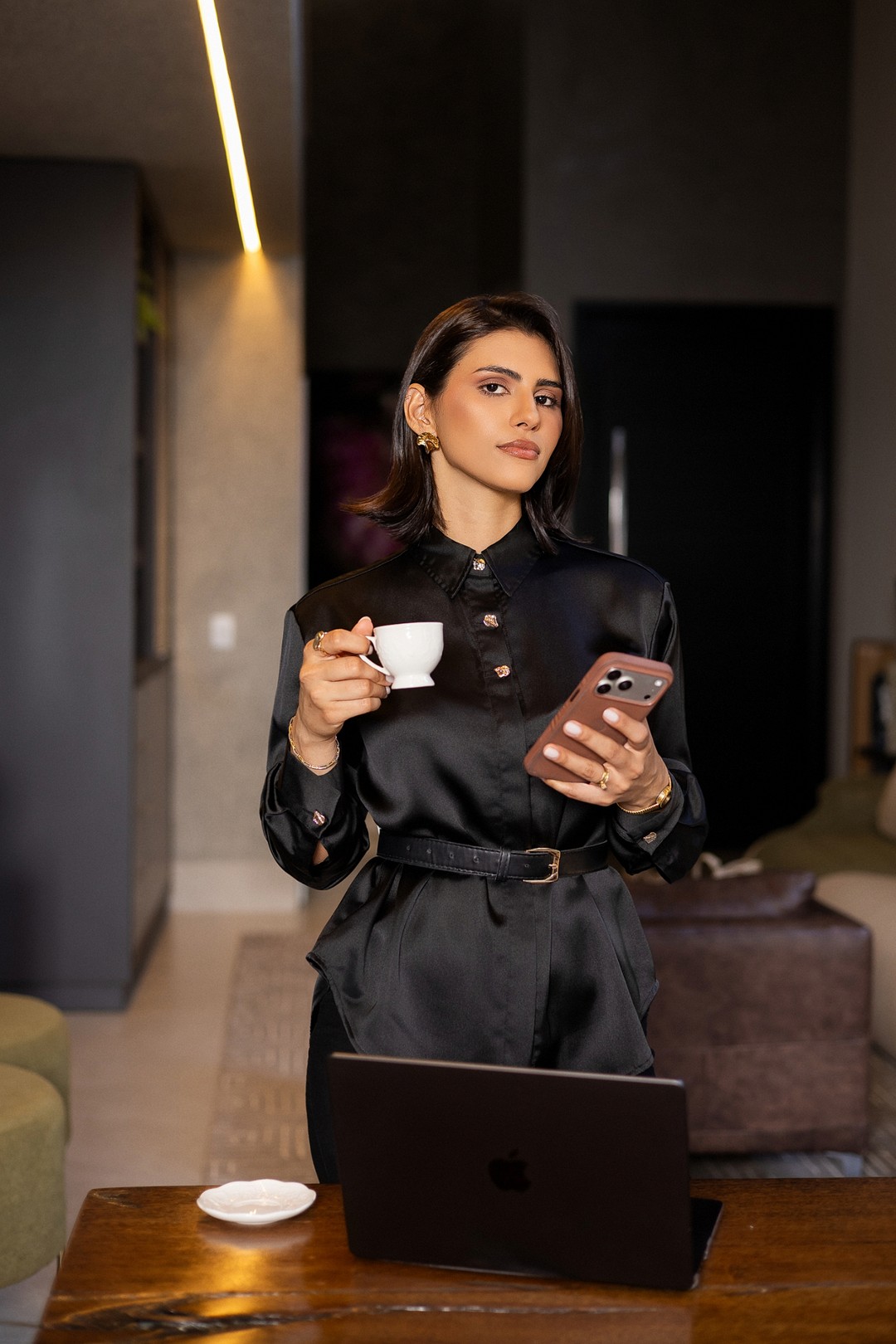 Yasmin de camisa de cetim preta, com uma xícara de café e o celular na mão, olhando para a câmera